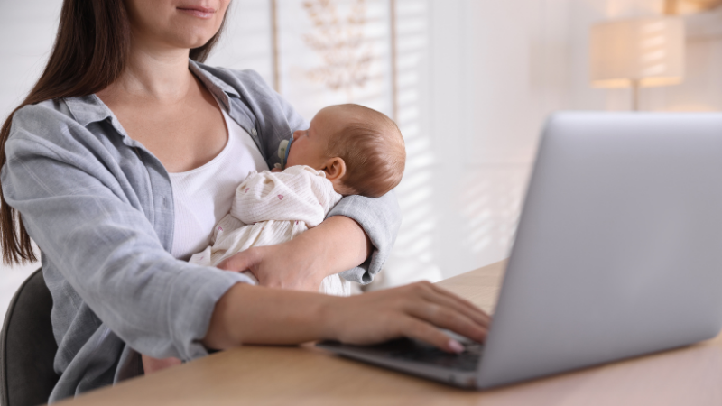 Person holding an infant while using a laptop during a telehealth healthcare appointment at home.